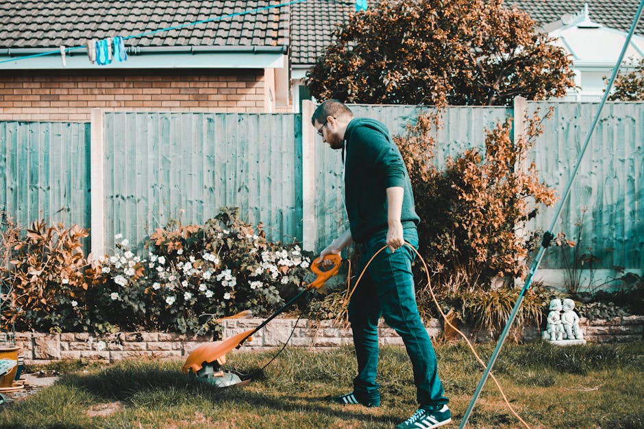 A man wearing a dark hoodie, blue jeans, and striped sneakers is trimming the grass with an electric lawnmower in a residential garden. The garden features a well-maintained lawn bordered by flower be